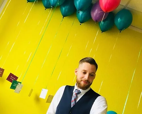 Mann, der vor einer gelben Wand steht, hinter ihm fliegen Luftballons mit Lernkarten.